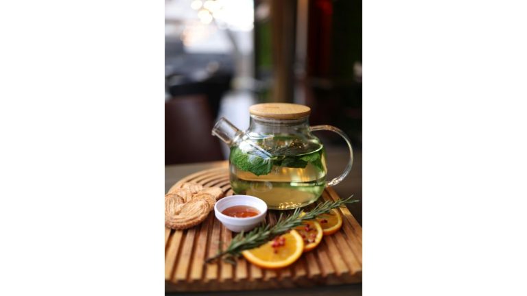 Théière en verre contenant une infusion, avec des agrumes et une branche de romarin sur un plateau