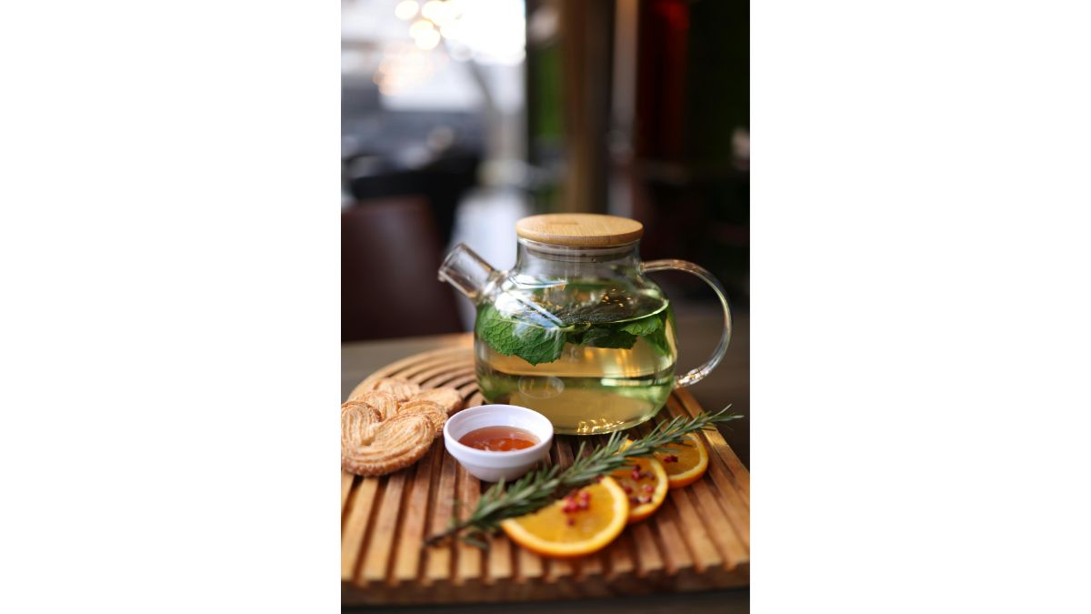Théière en verre contenant une infusion, avec des agrumes et une branche de romarin sur un plateau